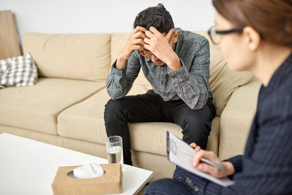 Fathers in recovery expressing emotions in therapy, man with head in hands while counselor listens and takes notes.